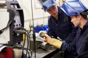 Two industrial workers performing welding and fabrication on metal components in a workshop setting with protective gear and equipment