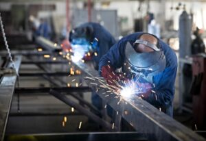 Two workers welding metal beams in an industrial workshop, wearing protective gear and helmets with sparks flying during fabrication process.