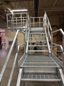 Galvanized steel truck washing staircase with expanded-metal steps and dual swing platforms in an indoor assembly area.