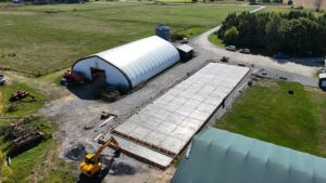 Aerial view of a concrete pad installation underway beside two fabric-covered agricultural buildings on a rural property.