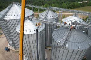 Workers performing installation and maintenance on large grain silos and elevated catwalk conveyors at an industrial storage facility.
