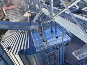 Custom engineered platform installed beside a grain bin, showing structural steel supports, guardrails, and overhead conveyor system.
