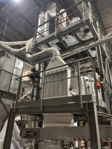 Industrial bulk material handling system featuring ductwork, mechanical equipment, steel support structure, and elevated service platforms inside a manufacturing facility.